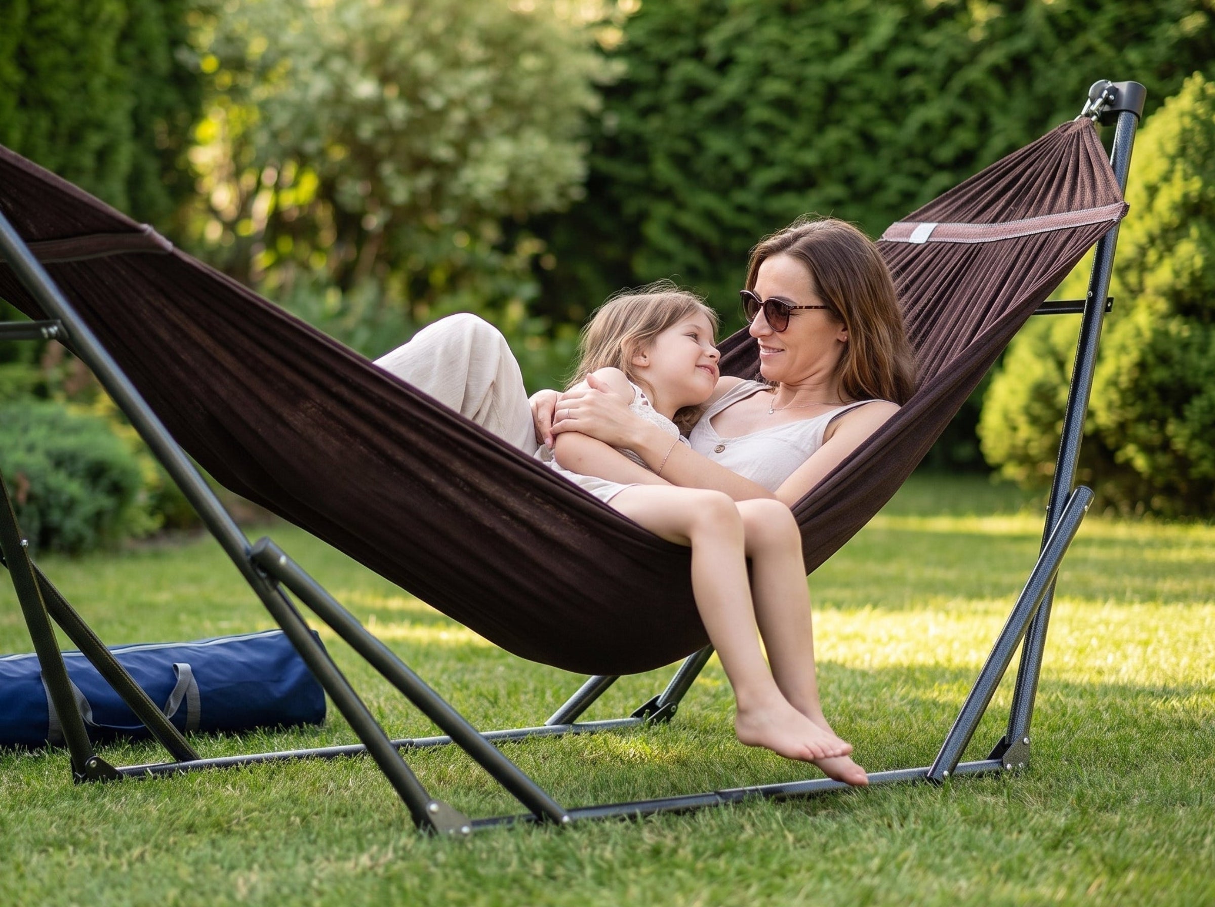 A woman and a child relax in a dark brown fabric hammock suspended on a black metal stand in a sunny green backyard. A blue carrying bag is placed on the grass beside the stand.
