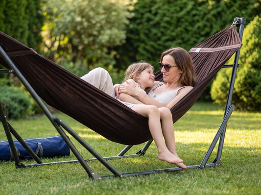 A woman and a child relax in a dark brown fabric hammock suspended on a black metal stand in a sunny green backyard. A blue carrying bag is placed on the grass beside the stand.